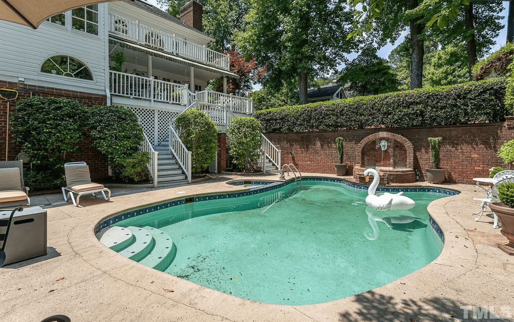 A light teal swimming pool with a floating swan, large privacy wall and hedge, and twin staircases leading to a back deck on a two-story white home